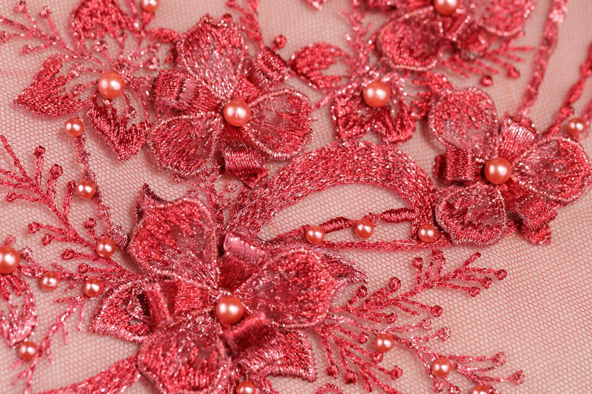 The image is a close-up of a delicate, intricate piece of red embroidery featuring a floral pattern with red flowers and small pearls.