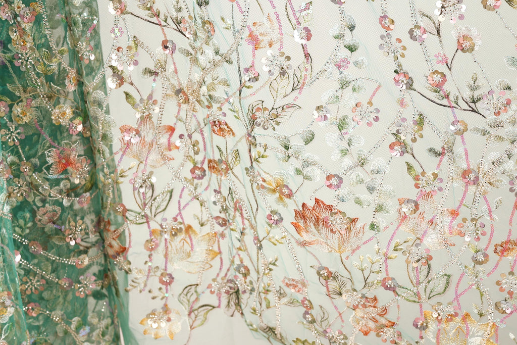 a close-up view of a fabric with a floral pattern, showcasing a variety of colorful flowers and leaves. The fabric appears to be made of a light-colored material, possibly cotton or linen, and is adorned with numerous small, colorful beads or sequins that add texture and visual interest to the design. The intricate details and vibrant colors of the floral pattern make the fabric a visually appealing and eye-catching piece.