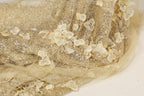 a close-up view of a delicate, shimmering fabric with a floral pattern. The fabric appears to be made of sheer material, giving it a delicate and ethereal appearance. The intricate design of the fabric features small, white flowers or leaves, which are the main decorative elements. The fabric is spread out on a surface, allowing for a clear and detailed view of its texture and pattern.