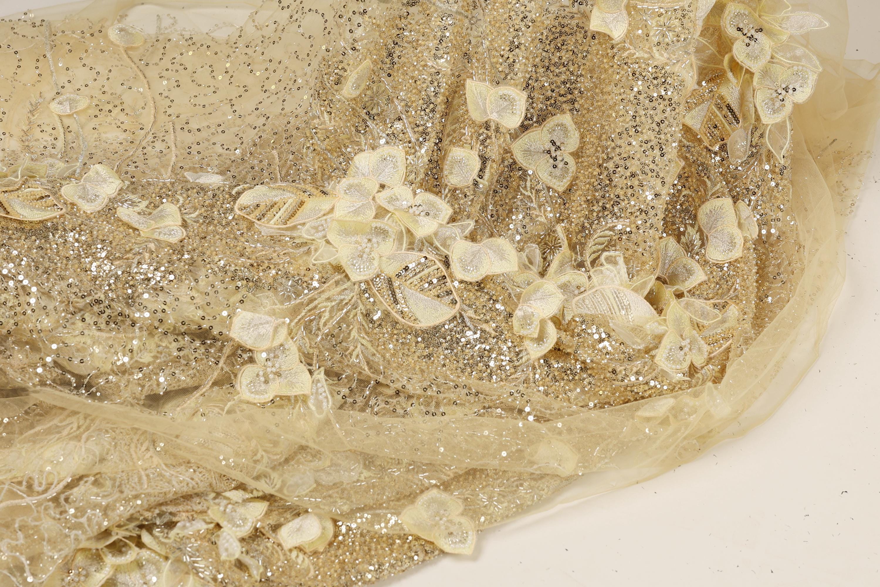 a close-up view of a delicate, shimmering fabric with a floral pattern. The fabric appears to be made of sheer material, giving it a delicate and ethereal appearance. The intricate design of the fabric features small, white flowers or leaves, which are the main decorative elements. The fabric is spread out on a surface, allowing for a clear and detailed view of its texture and pattern.
