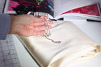 A hand holding a piece of jewelry, possibly a necklace or bracelet, is resting on a white fabric surface. In the background, there are several books and a pair of scissors, suggesting a workspace or study area.