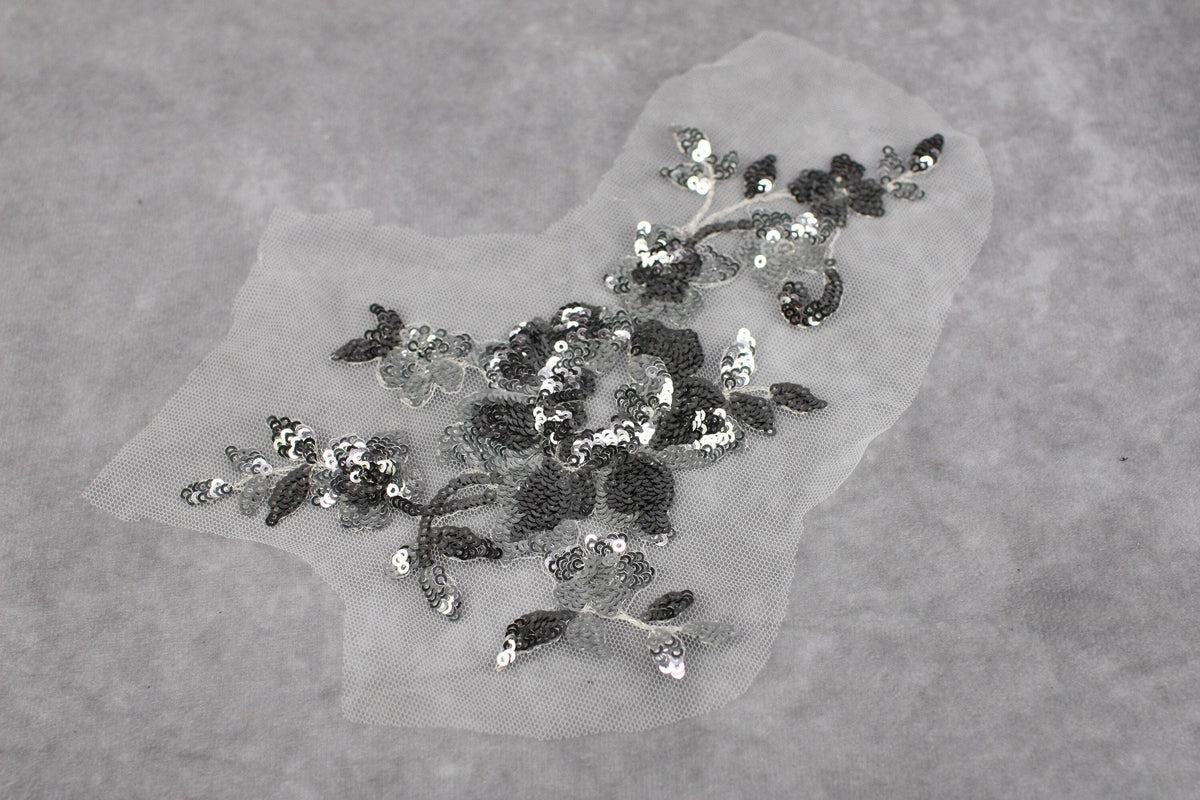 a close-up view of a delicate, intricate piece of fabric with a floral pattern. The fabric appears to be made of a sheer material, giving it a delicate and ethereal appearance. The pattern consists of small, black and silver sequins that are meticulously arranged to create the floral design. The image does not contain any text or other discernible objects, and the focus is solely on the intricate details of the fabric.