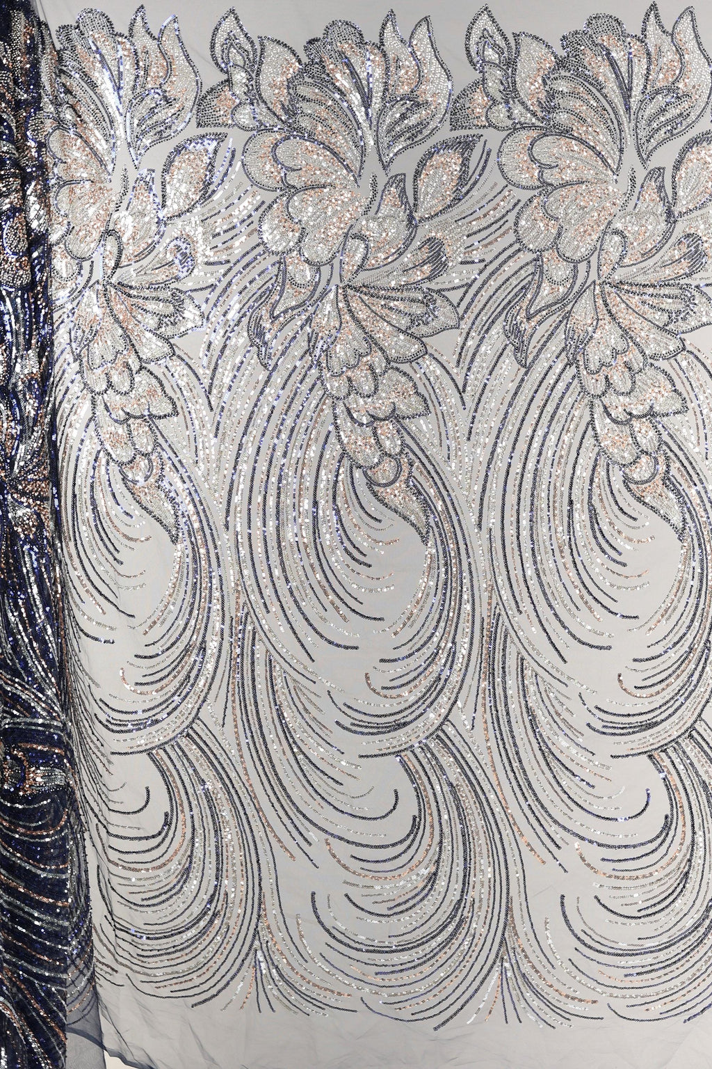 a close-up view of a fabric with a repeating pattern of silver and black sequin designs. The pattern consists of various shapes and swirls, creating a visually striking and intricate design. The fabric appears to be a light-colored material, possibly cotton or linen, with the silver and black sequins adding a touch of sparkle and elegance to the overall appearance.