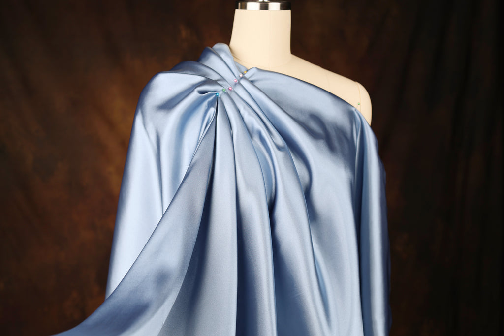 A mannequin wearing a blue satin one-shoulder dress with a draped, flowing silhouette.
