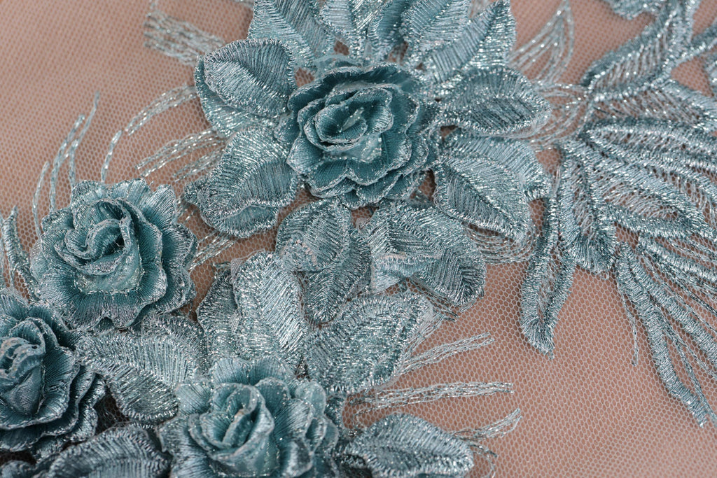 a close-up view of a delicate, intricately designed fabric with a floral pattern. The fabric features a variety of blue and green flowers, including roses and leaves, arranged in a visually appealing manner. The close-up perspective allows for a detailed examination of the fabric's texture and the intricate details of the floral design.