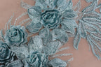 a close-up view of a delicate, intricately designed fabric with a floral pattern. The fabric features a variety of blue and green flowers, including roses and leaves, arranged in a visually appealing manner. The close-up perspective allows for a detailed examination of the fabric's texture and the intricate details of the floral design.