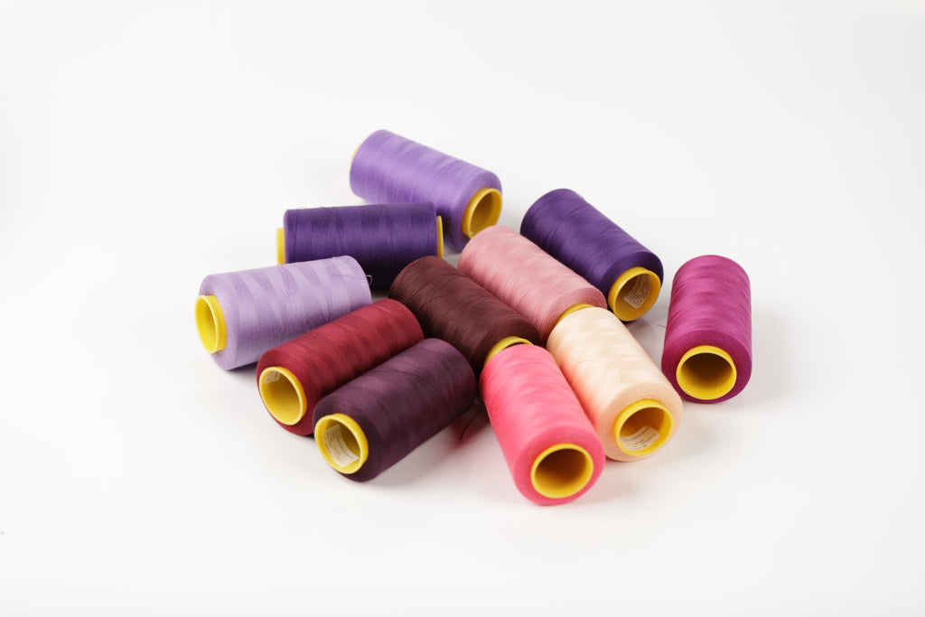 A collection of colorful spools of thread in various shades of purple, pink, and yellow are scattered on a white background.