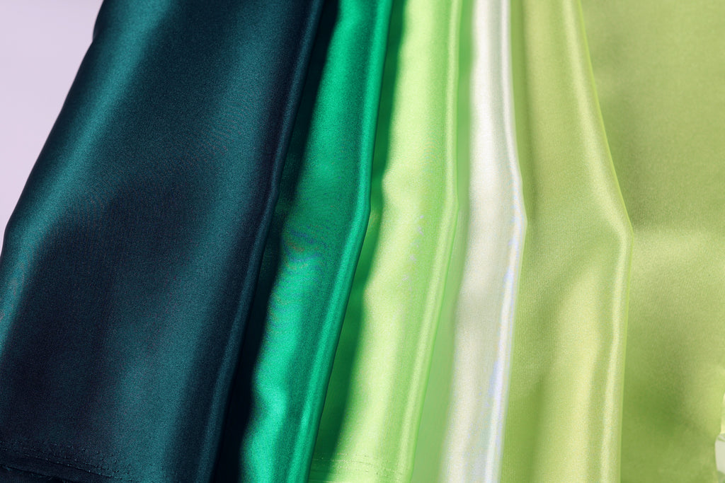 A close-up image of various colored satin fabric, including dark green, light green, and white, arranged in a vertical line.