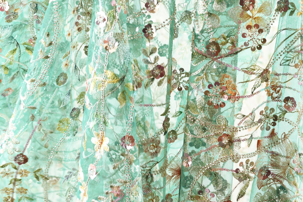 The image is a close-up of a fabric with a floral pattern, featuring various shades of green, pink, and yellow flowers and leaves. The fabric appears to be made of a sheer material, giving it a delicate and airy appearance.