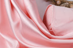 A close-up view of a soft, flowing pink satin fabric, with a blurred background that appears to be a plant or floral arrangement.