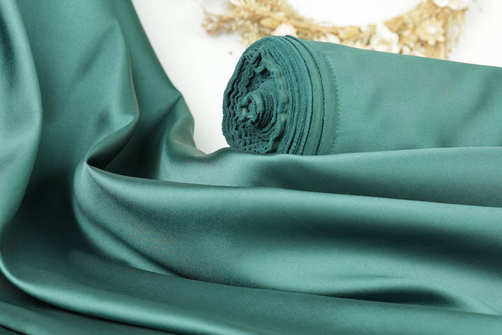 A close-up view of a green satin fabric, possibly silk, with a rolled-up end visible. The fabric appears to be smooth and shiny, suggesting it is made of a high-quality material.