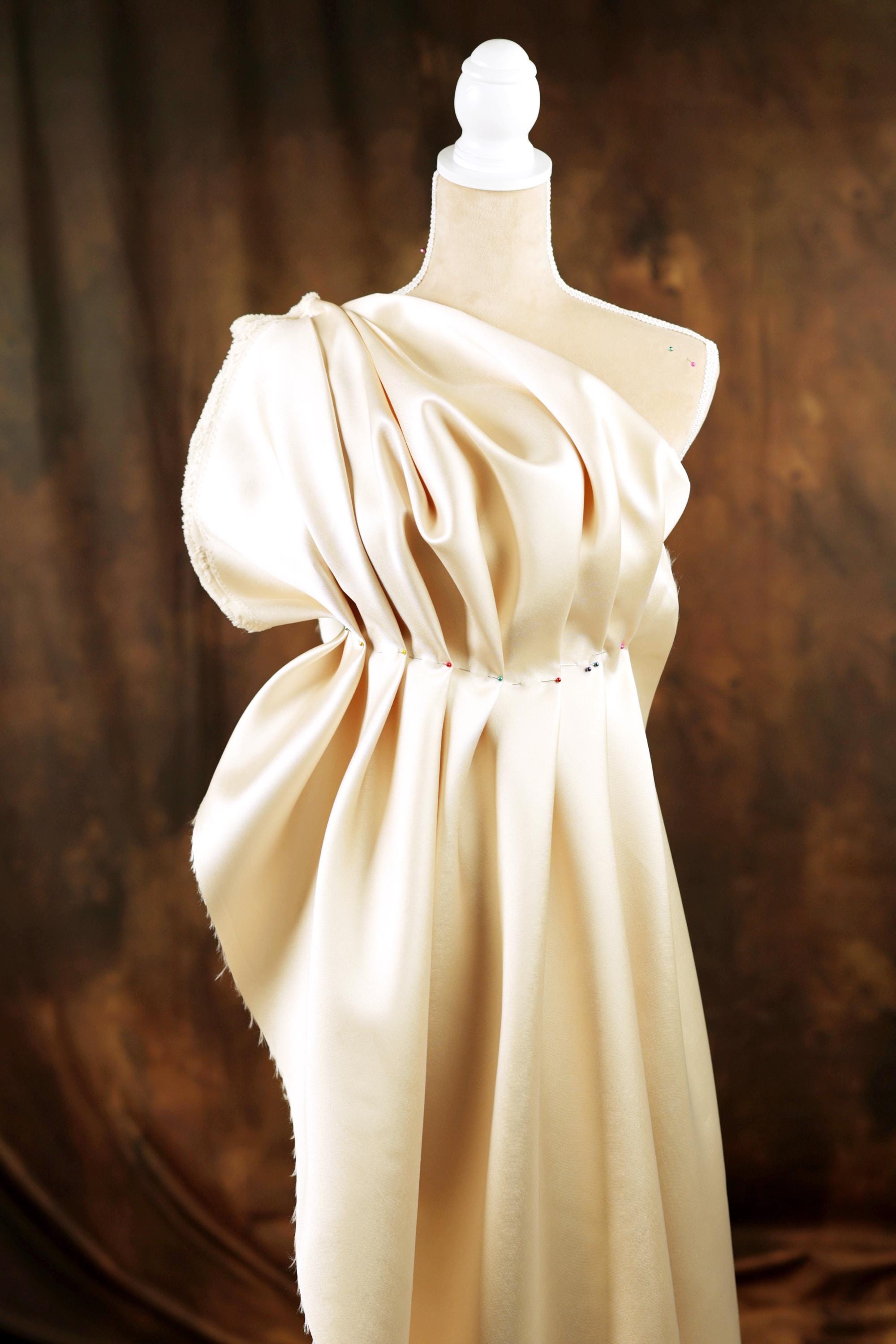 A white mannequin wearing a flowing, draped white dress against a brown background.