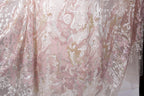 a close-up view of a delicate, lace-like fabric with a floral pattern in shades of pink and white. The fabric appears to be made of sheer material, giving it a delicate and airy appearance.