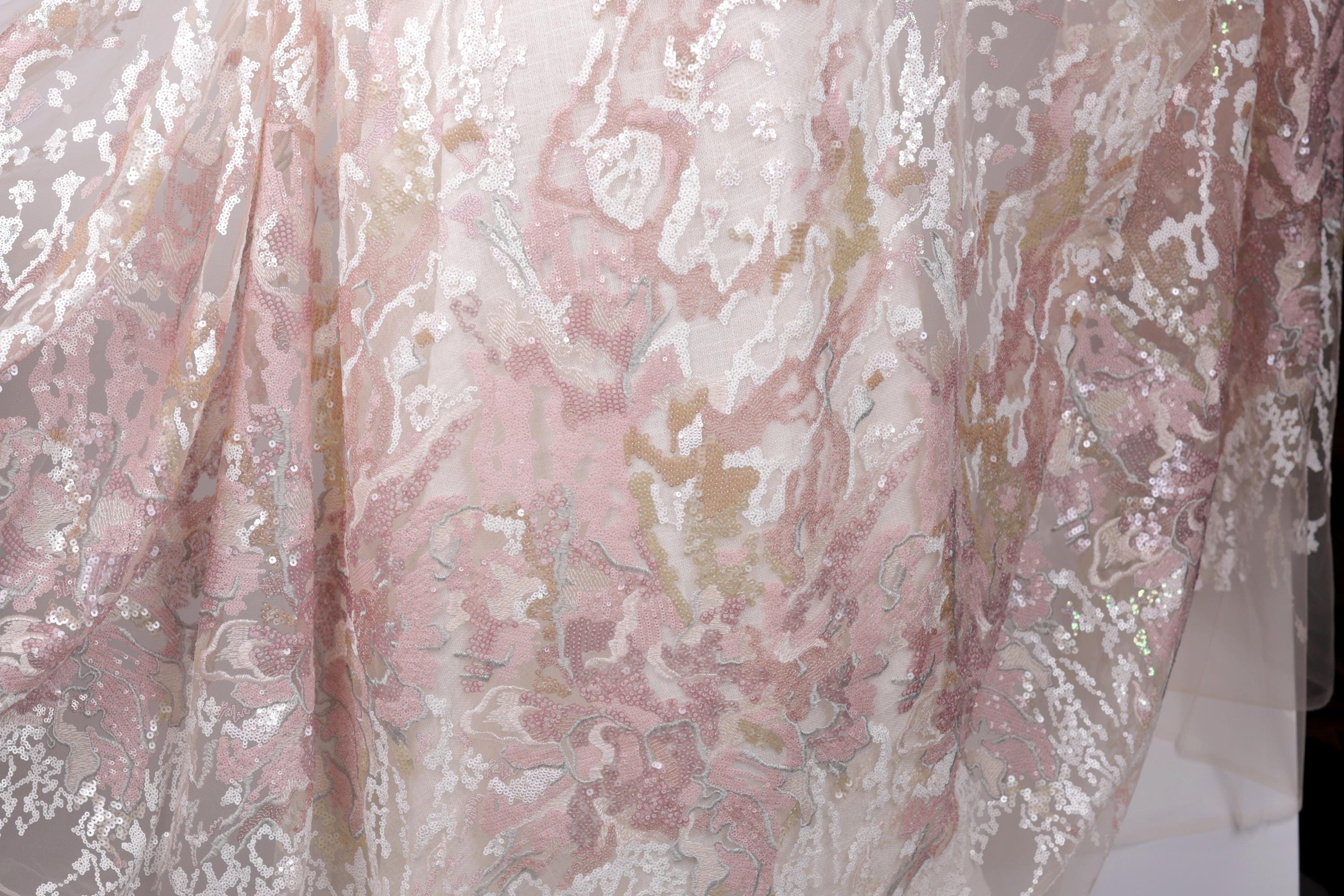 a close-up view of a delicate, lace-like fabric with a floral pattern in shades of pink and white. The fabric appears to be made of sheer material, giving it a delicate and airy appearance.