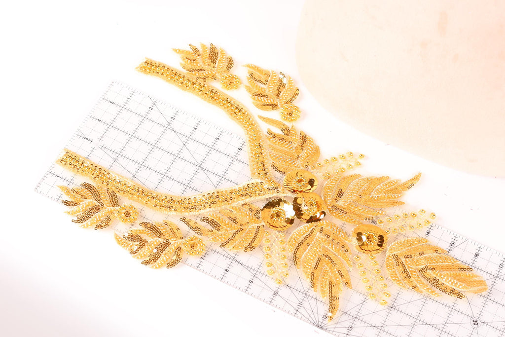 a close-up view of a gold-colored embroidered piece with intricate details, including leaves and flowers, placed on a white surface.