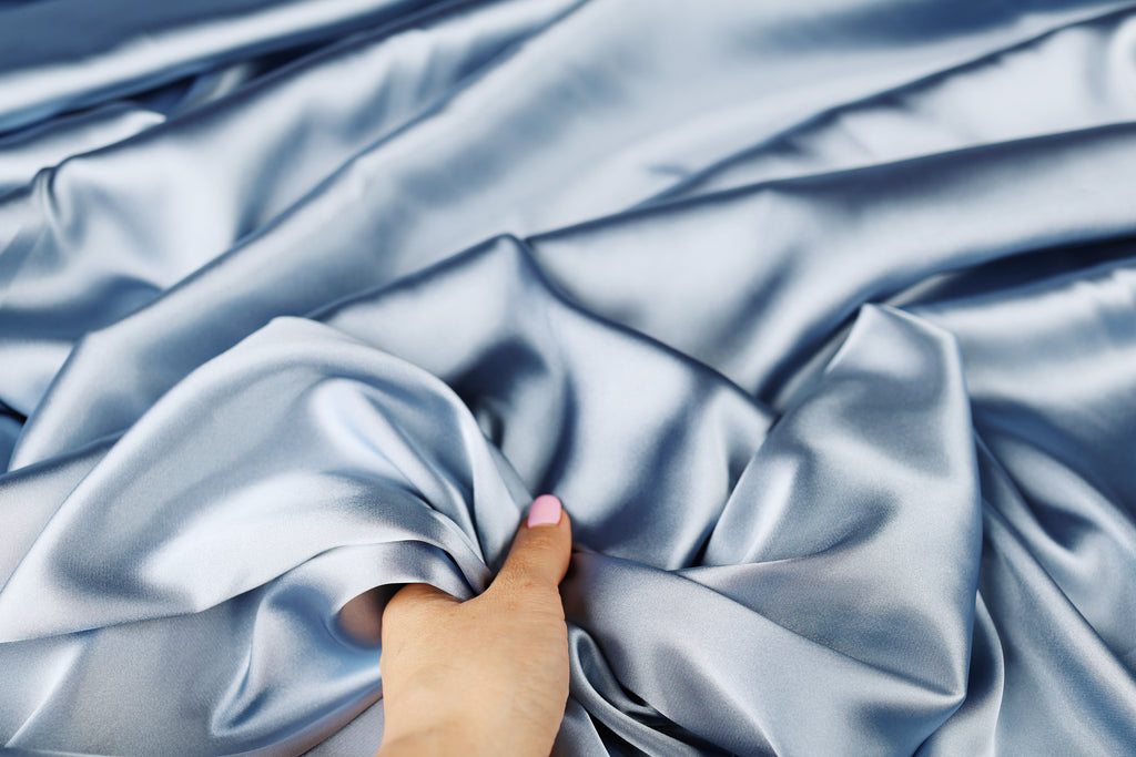 A hand is holding a piece of blue satin fabric, which appears to be soft and smooth.