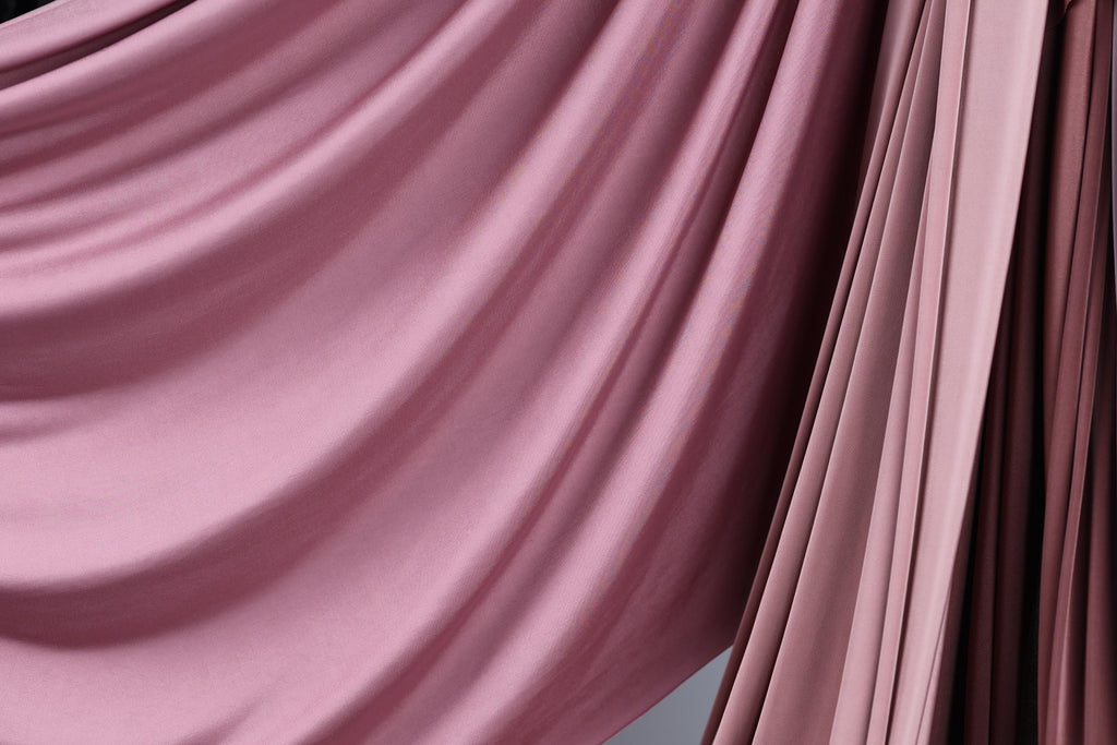 a close-up view of a flowing, draped fabric with a soft, pinkish hue. The fabric appears to be made of a smooth, shiny material, possibly silk or satin, and is draped in a way that creates a sense of movement and elegance. The fabric is draped over a surface, adding a touch of sophistication and style to the scene.