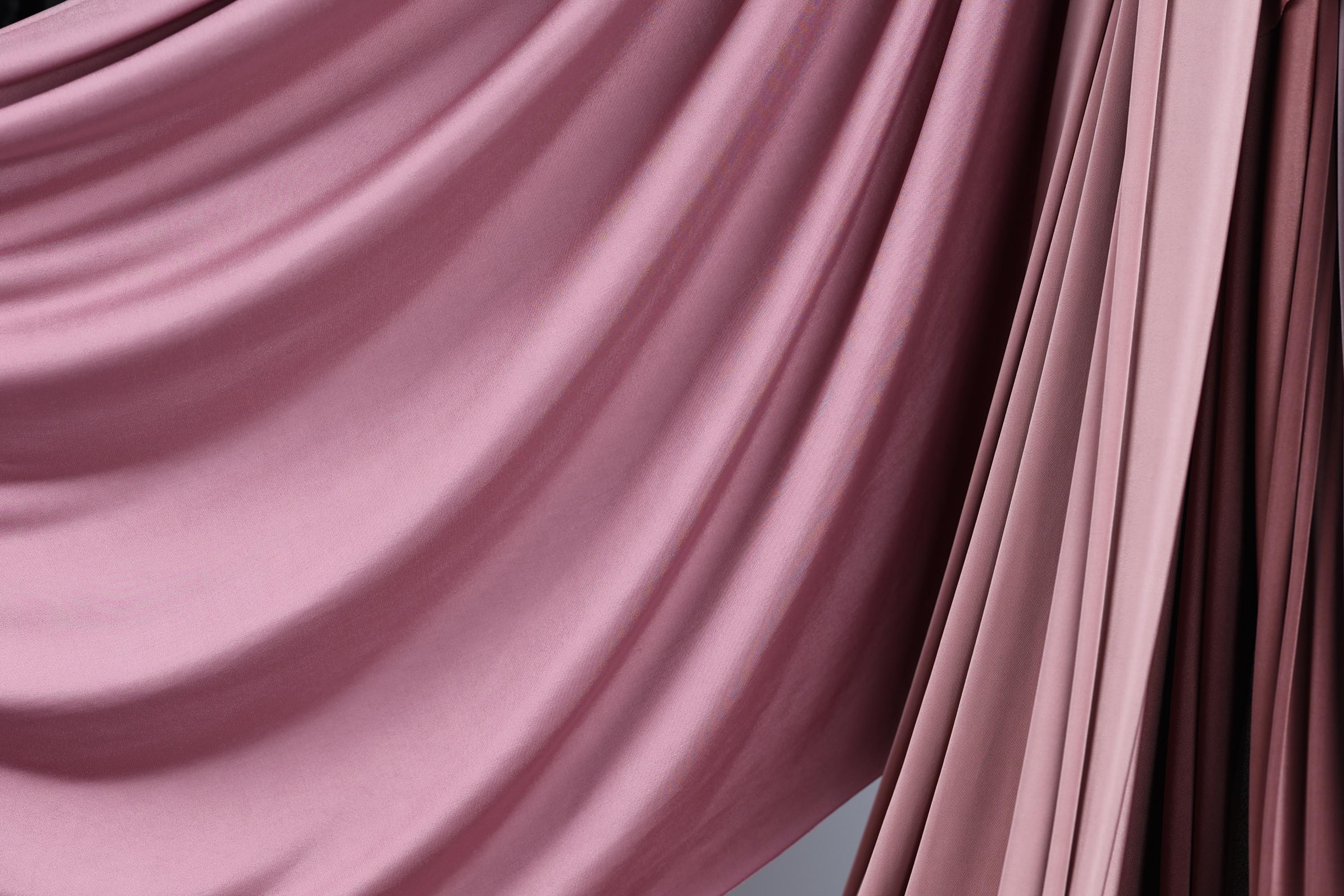 a close-up view of a flowing, draped fabric with a soft, pinkish hue. The fabric appears to be made of a smooth, shiny material, possibly silk or satin, and is draped in a way that creates a sense of movement and elegance. The fabric is draped over a surface, adding a touch of sophistication and style to the scene.