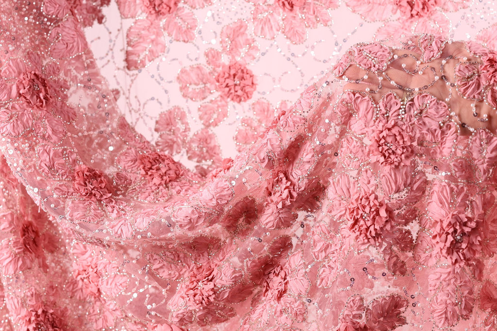 The image is a close-up of a delicate, pink lace fabric with intricate floral patterns. The fabric appears to be made of sheer material, giving it a soft and delicate appearance. The lace is adorned with numerous small flowers, creating a visually appealing and intricate design.
