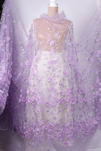 A mannequin wearing a white dress with a sheer, floral-patterned fabric draped over it. The dress has a long, flowing skirt and a high neckline, creating a romantic and elegant appearance.