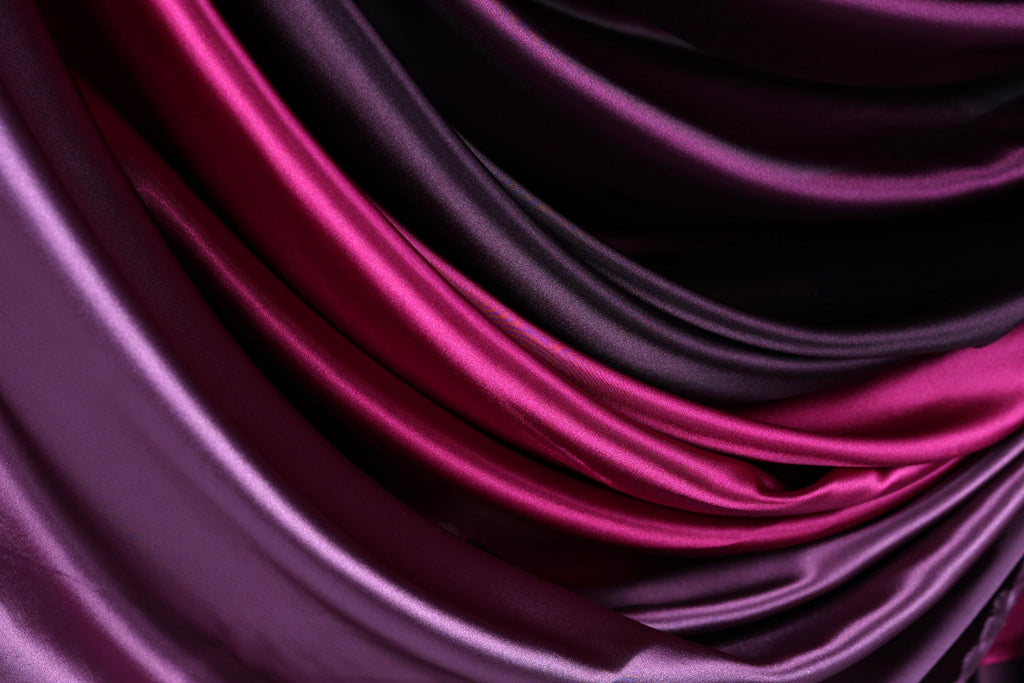 a close-up view of a fabric with a gradient of colors, transitioning from a deep purple to a lighter shade of purple. The fabric appears to be made of satin or a similar smooth, shiny material.