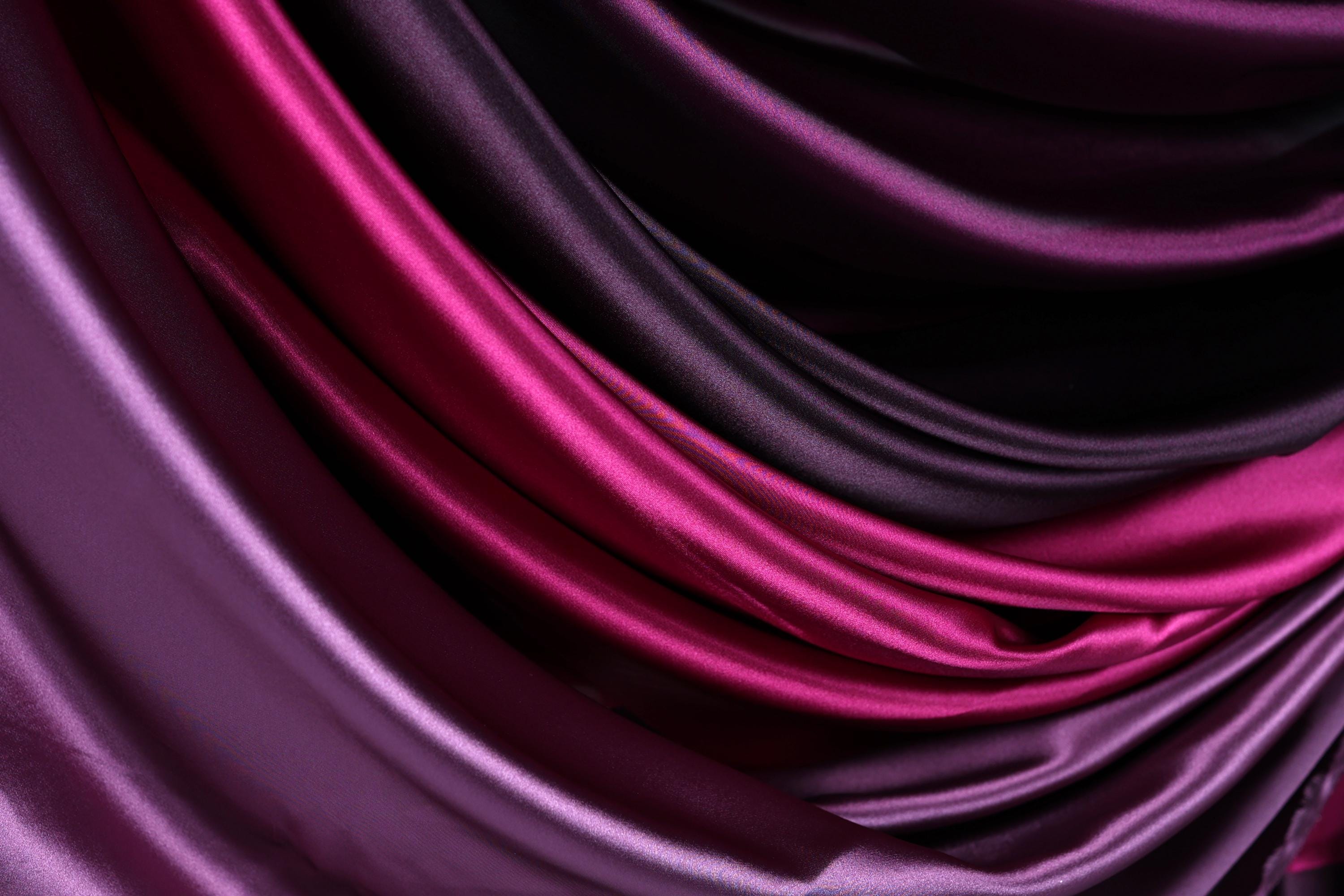 a close-up view of a fabric with a gradient of colors, transitioning from a deep purple to a lighter shade of purple. The fabric appears to be made of satin or a similar smooth, shiny material.