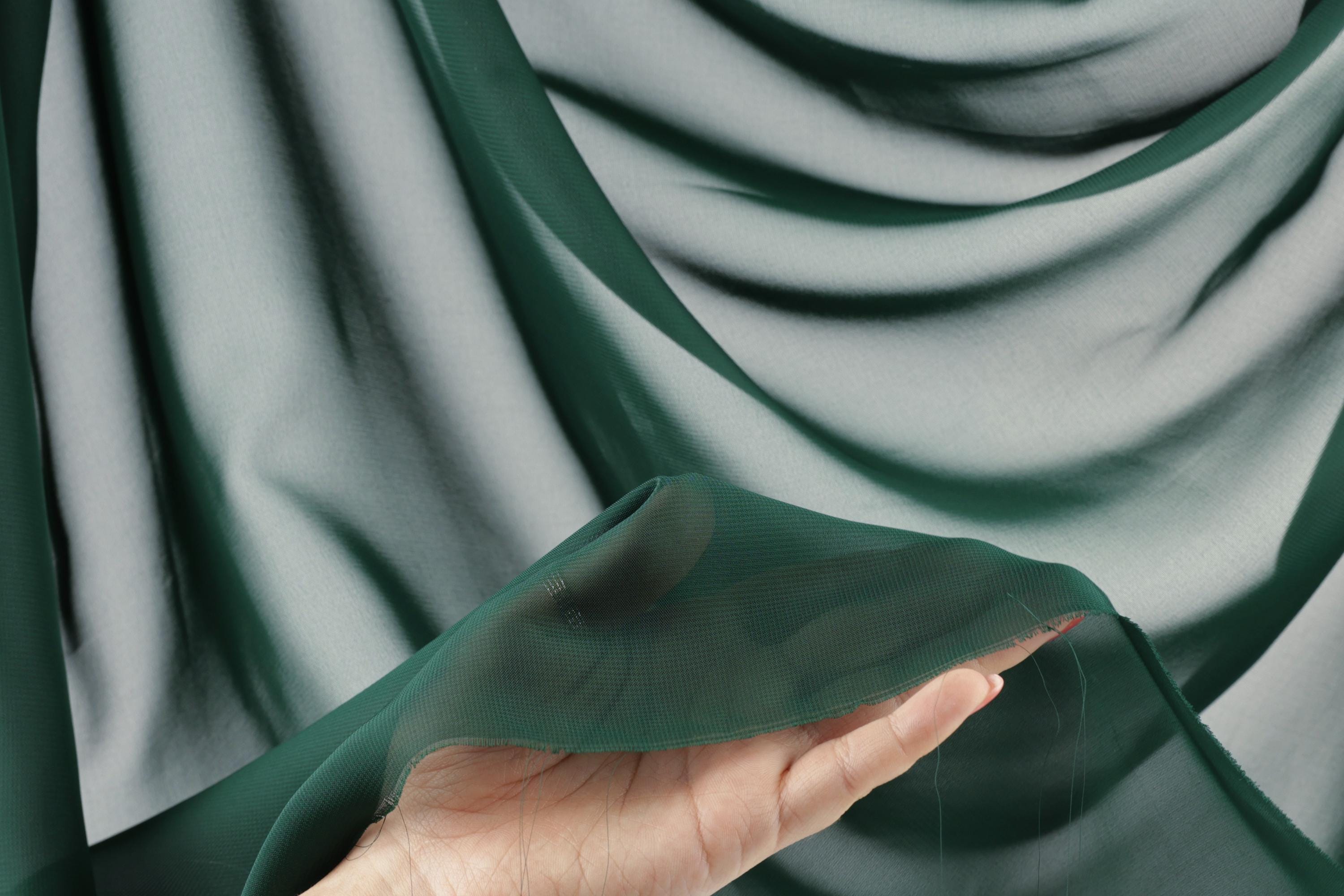 A hand is holding a piece of green fabric against a blurred background of green fabric.