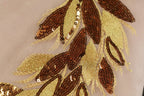a close-up view of a delicate, intricate embroidered design with a combination of brown and gold sequins and beads. The design appears to be a floral pattern, with the individual elements meticulously arranged to create a visually appealing and detailed piece of textile art.