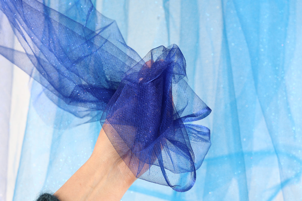 A hand is holding a piece of blue, glittery fabric against a blue background.