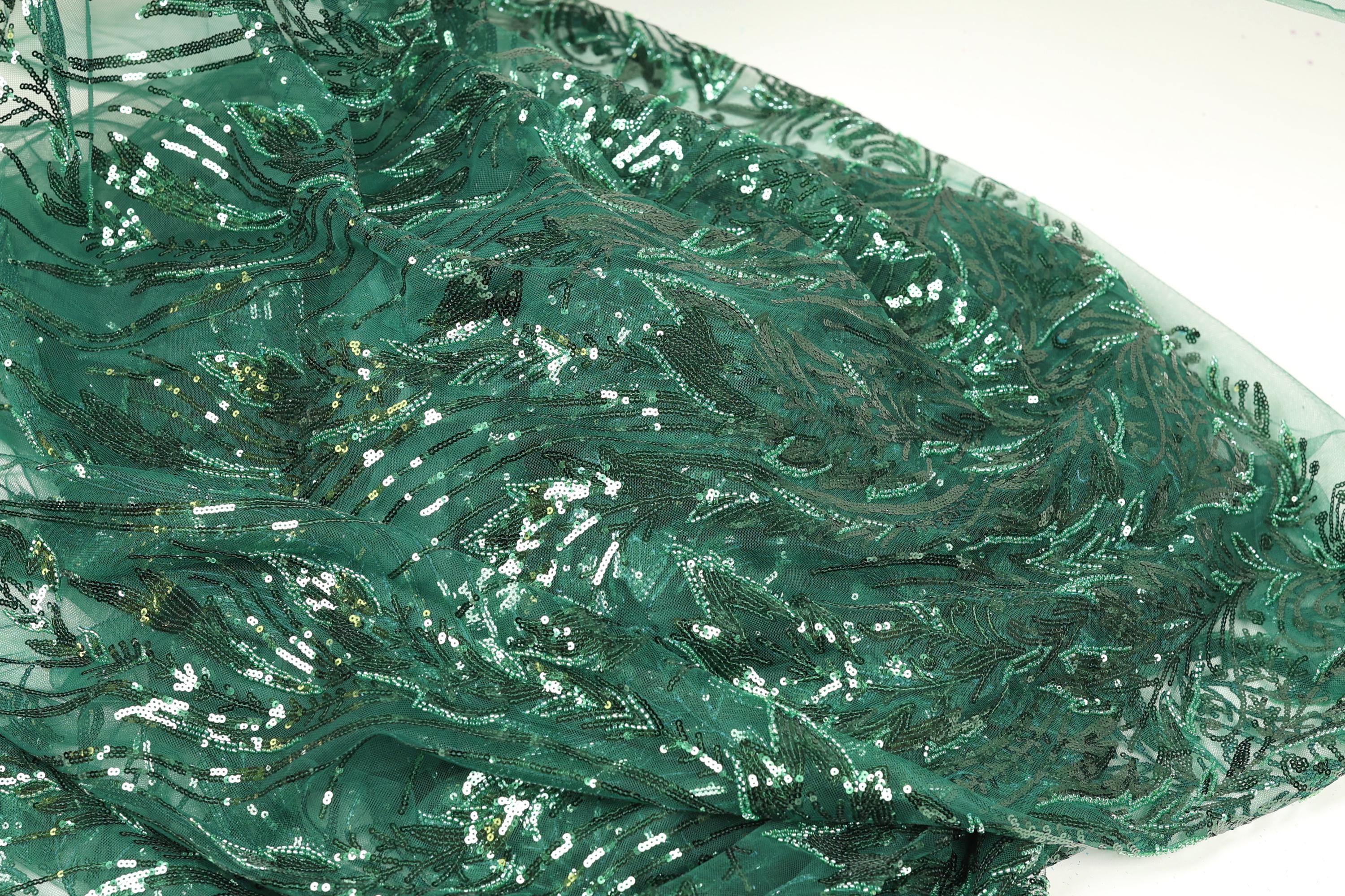 a close-up view of a fabric with a green, sequined pattern. The fabric appears to be made of a shiny, green material with a floral or leaf-like design. The fabric is folded and crumpled, giving it a textured appearance.