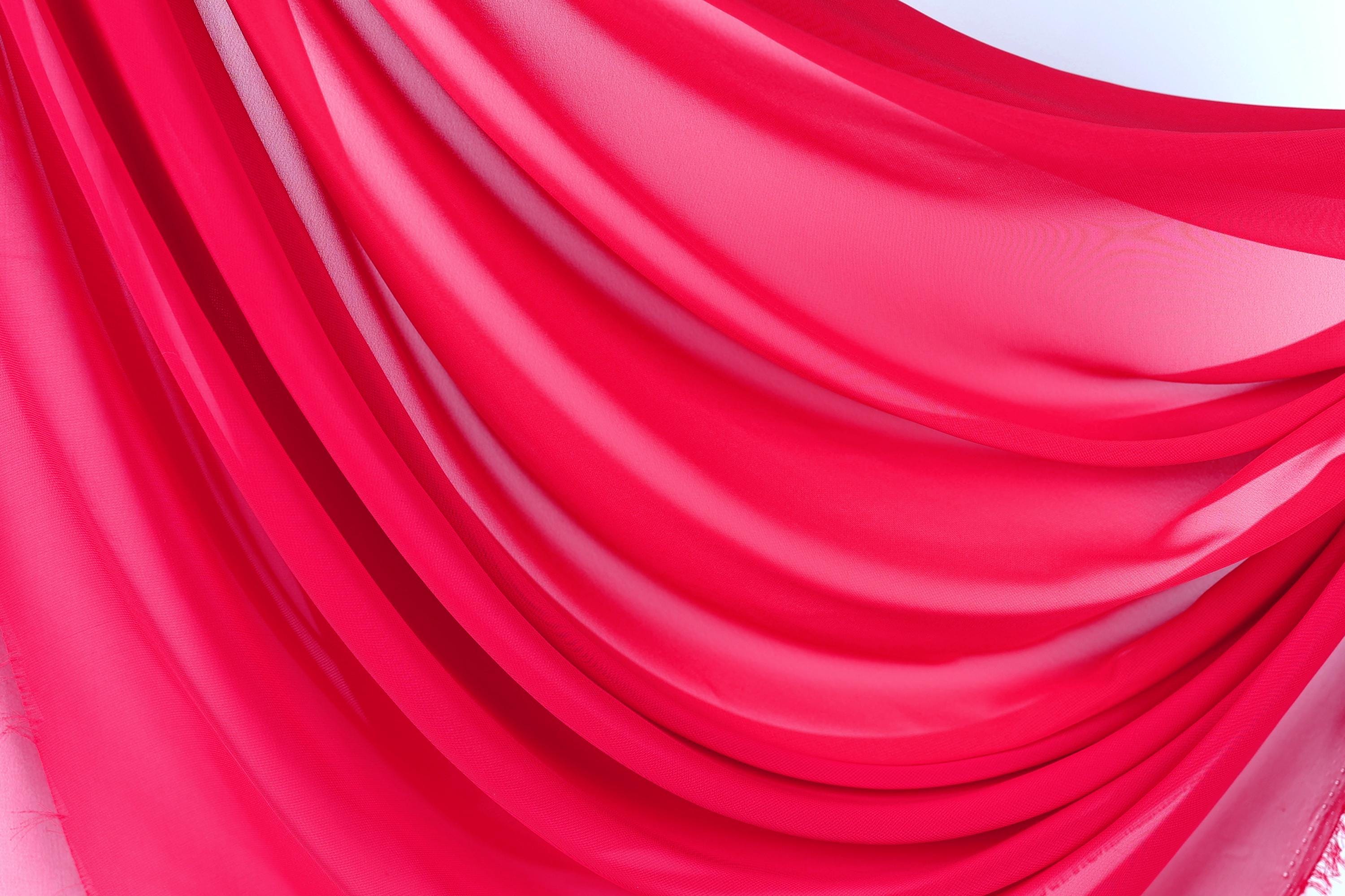 The image is a close-up of a vibrant red fabric, possibly a chiffon or sheer material, with a smooth, flowing texture. The fabric appears to be draped or draped over an object, creating a sense of movement and softness.