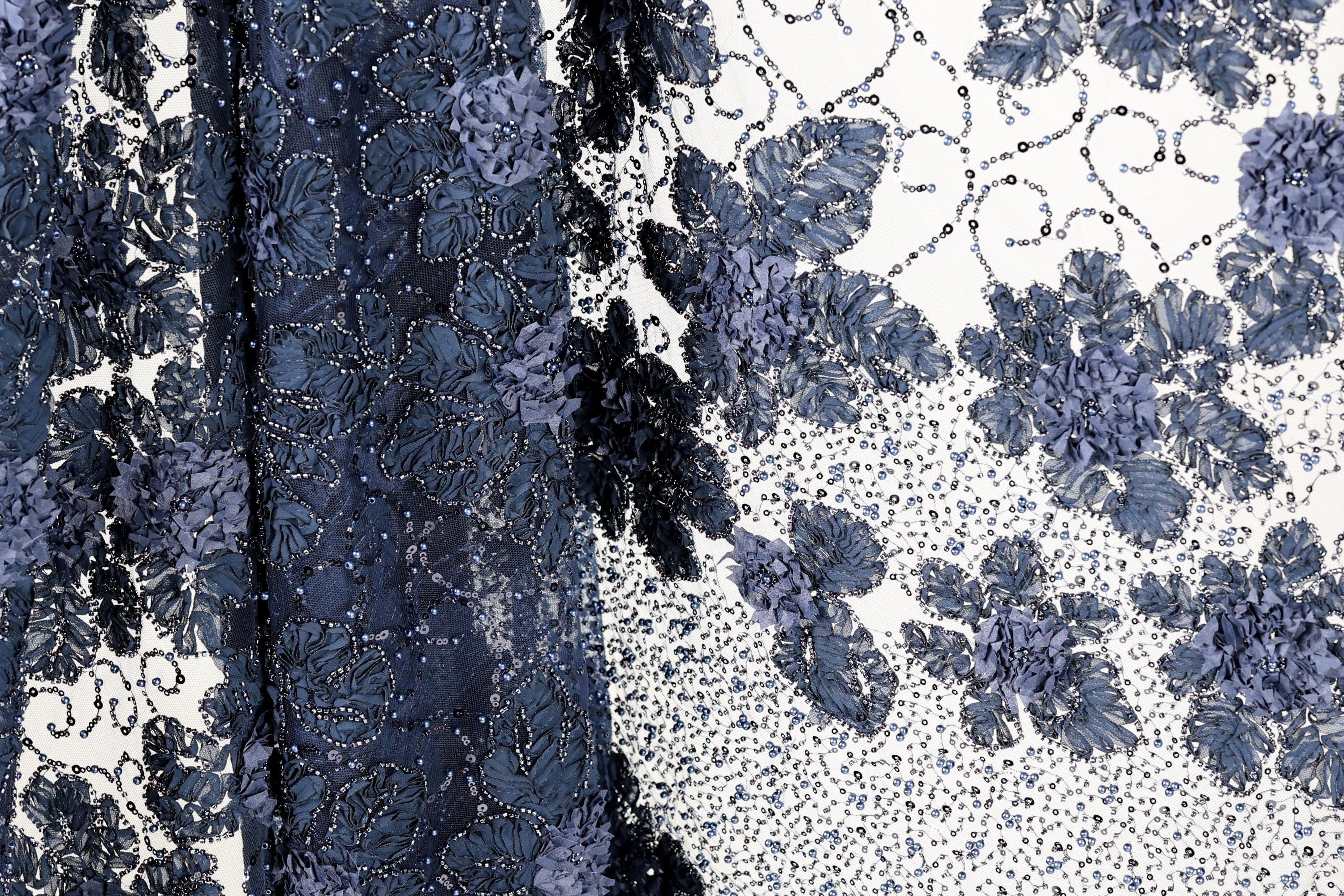 a close-up view of a fabric with a floral pattern, including blue flowers and leaves. The fabric appears to be made of a material with a textured surface, possibly lace or a similar type of material. The image does not contain any text or other discernible objects or actions.