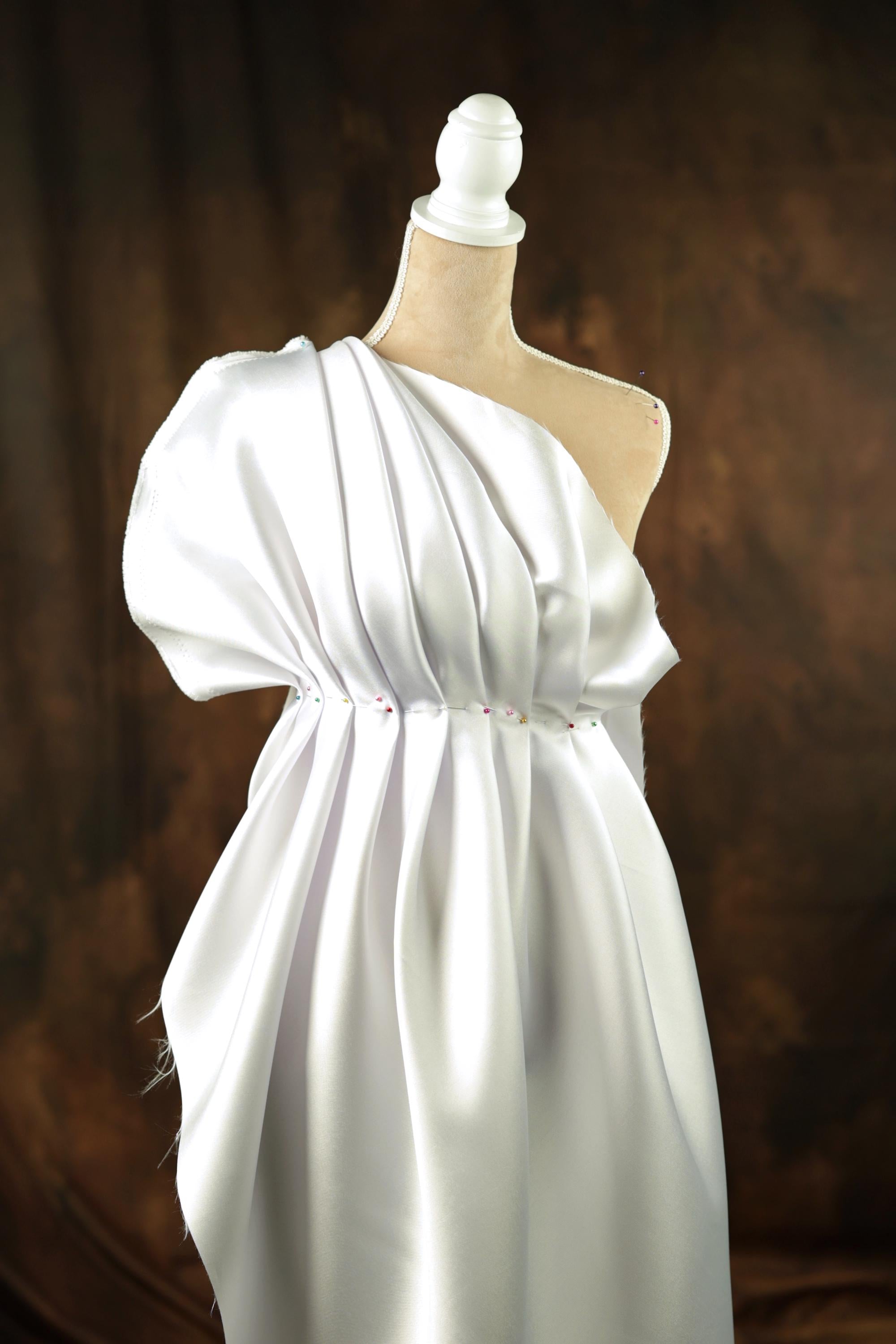 A white, one-shoulder dress displayed on a mannequin against a brown background.
