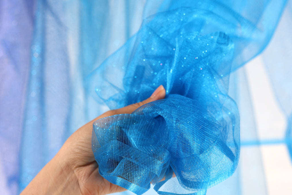 A hand is holding a piece of blue glittery fabric, which appears to be a piece of sheer material.