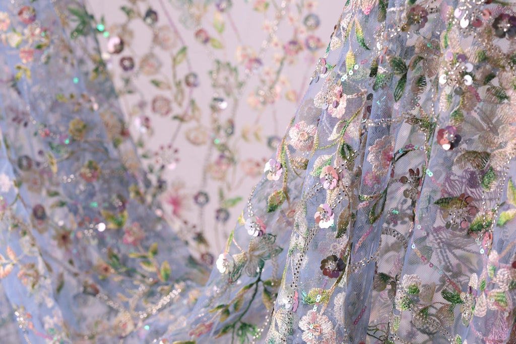 a close-up view of a delicate, intricately embroidered fabric with a floral pattern. The fabric appears to be made of a light blue material, and the embroidery is adorned with small, colorful flowers and leaves. The image is a close-up of the fabric, showcasing the detailed craftsmanship and the vibrant colors of the embroidery.