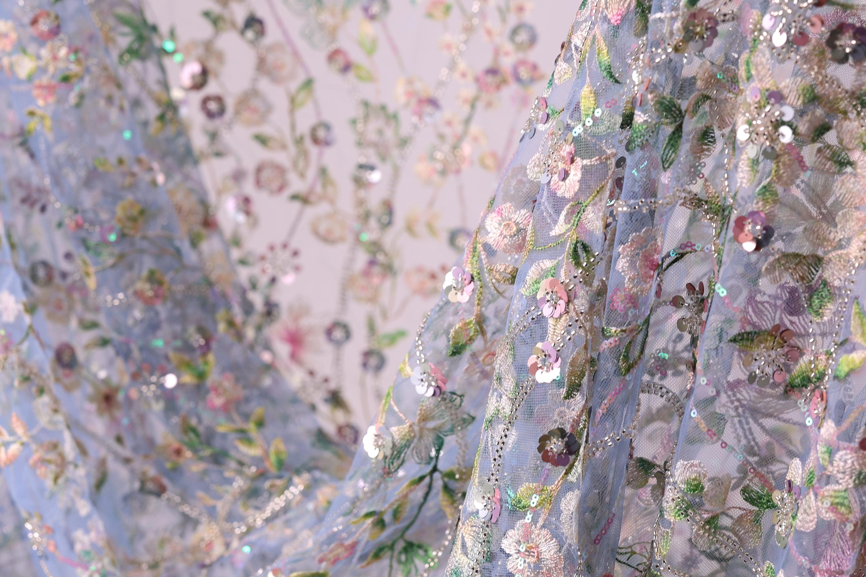 a close-up view of a delicate, intricately embroidered fabric with a floral pattern. The fabric appears to be made of a light blue material, and the embroidery is adorned with small, colorful flowers and leaves. The image is a close-up of the fabric, showcasing the detailed craftsmanship and the vibrant colors of the embroidery.