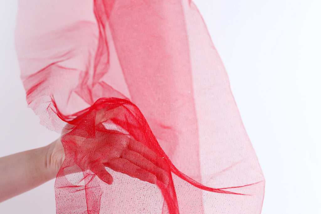 A hand is holding a red fabric, which appears to be a sheer or sheer-like material, against a white background.
