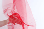 A hand is holding a red fabric, which appears to be a sheer or sheer-like material, against a white background.