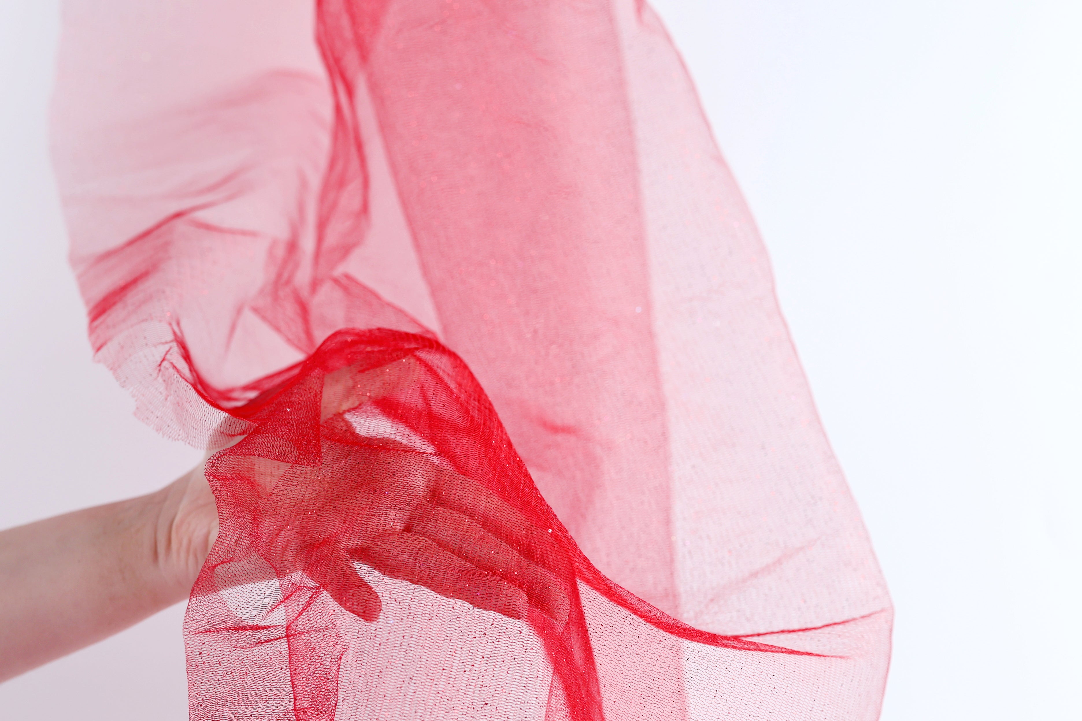 A hand is holding a red fabric, which appears to be a sheer or sheer-like material, against a white background.