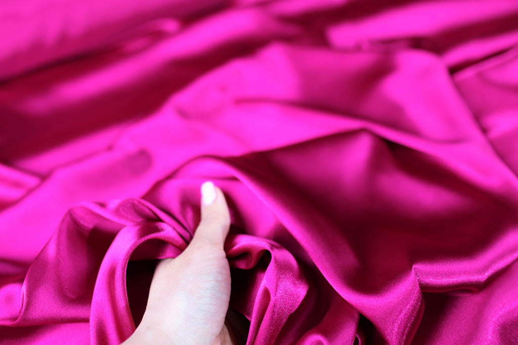 A hand is holding a piece of bright pink satin fabric, which appears to be soft and smooth.