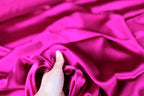 A hand is holding a piece of bright pink satin fabric, which appears to be soft and smooth.
