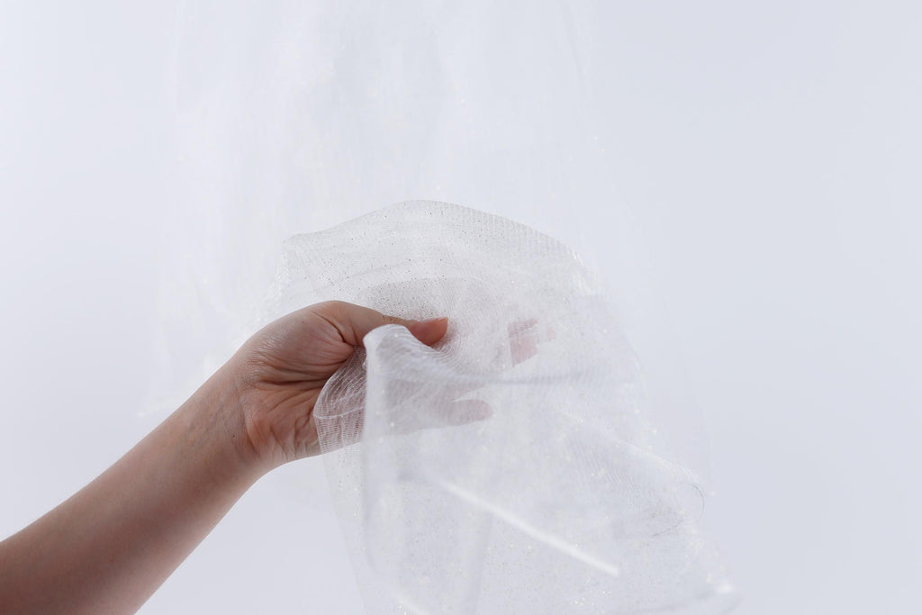 A hand is holding a piece of white fabric, possibly a veil or a sheer material, against a plain white background.
