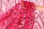 A hand is holding up a piece of pink sequin fabric against a pink background with a repeating pattern of pink and black sequins.