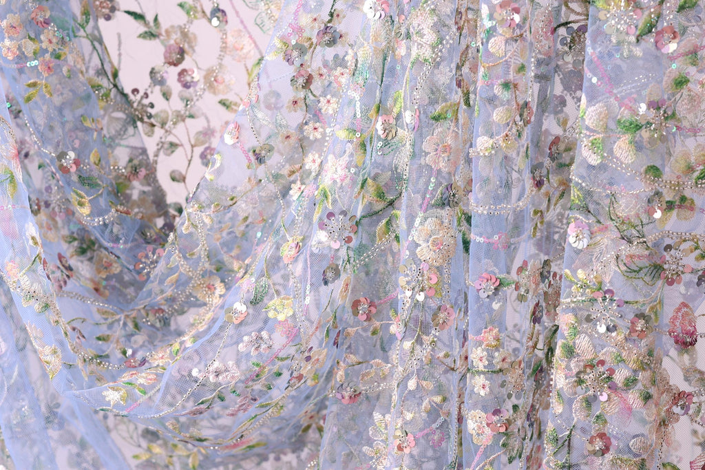 a close-up view of a delicate, intricately embroidered fabric with a floral pattern. The fabric appears to be made of sheer material, allowing the light to pass through and revealing the detailed embroidery work. The colors used in the design are predominantly pastel shades, creating a soft and delicate appearance.