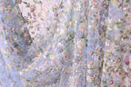 a close-up view of a delicate, intricately embroidered fabric with a floral pattern. The fabric appears to be made of sheer material, allowing the light to pass through and revealing the detailed embroidery work. The colors used in the design are predominantly pastel shades, creating a soft and delicate appearance.