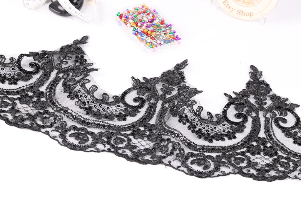 a close-up view of a black lace trim with intricate floral designs. The lace appears to be a decorative piece, possibly used for sewing or crafting purposes. In the background, there are various sewing and crafting supplies, such as a sewing kit and a box of colorful pins.
