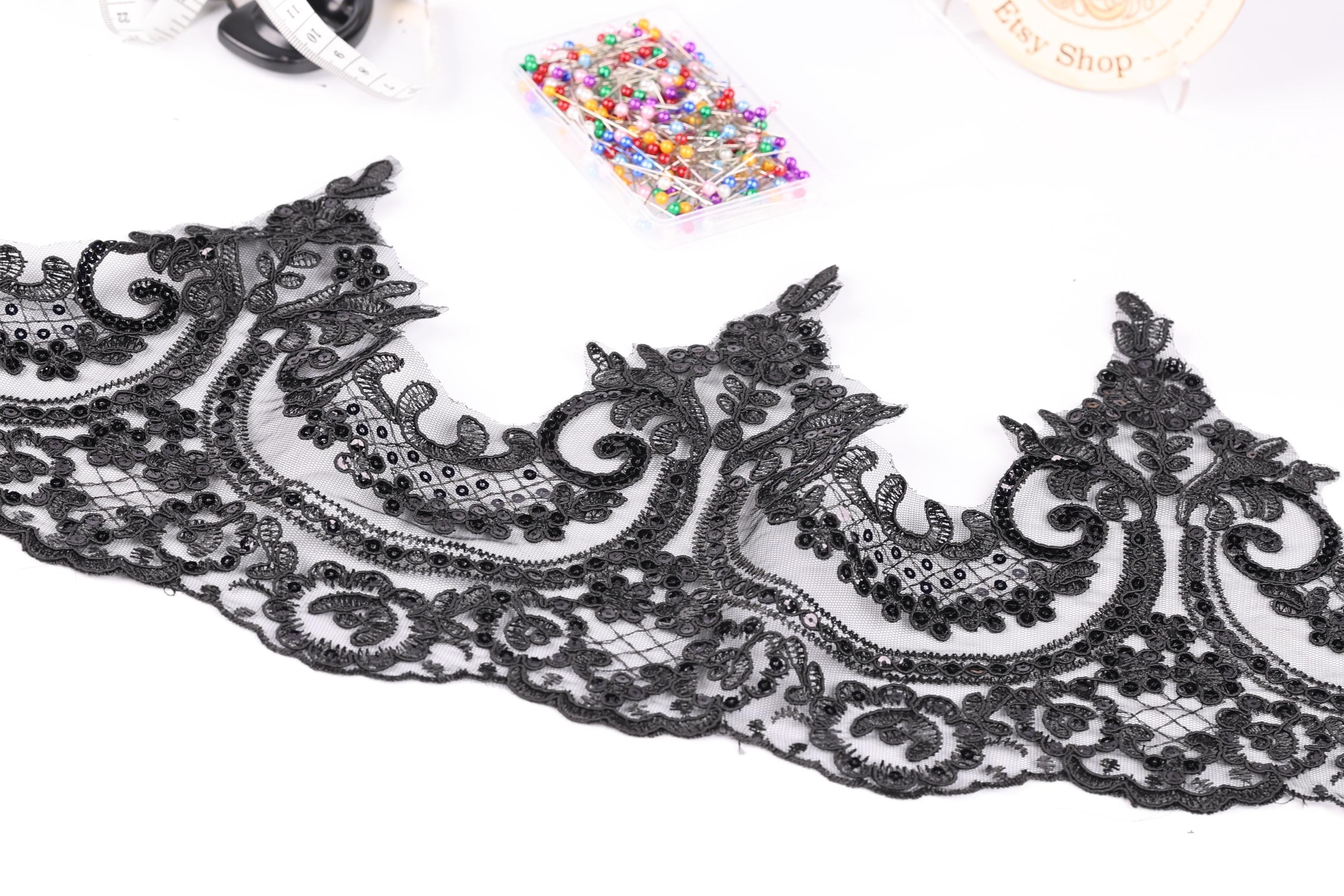 a close-up view of a black lace trim with intricate floral designs. The lace appears to be a decorative piece, possibly used for sewing or crafting purposes. In the background, there are various sewing and crafting supplies, such as a sewing kit and a box of colorful pins.