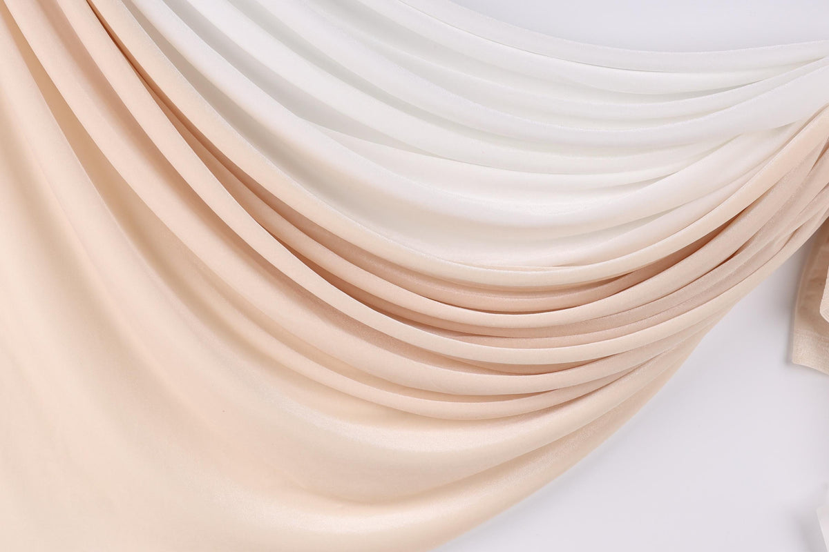 a close-up view of a fabric with a gradient of colors, transitioning from light to dark shades of peach and white. The fabric appears to be made of a soft, flowing material, possibly silk or chiffon, and is draped in a cascading manner, creating a sense of movement and elegance.