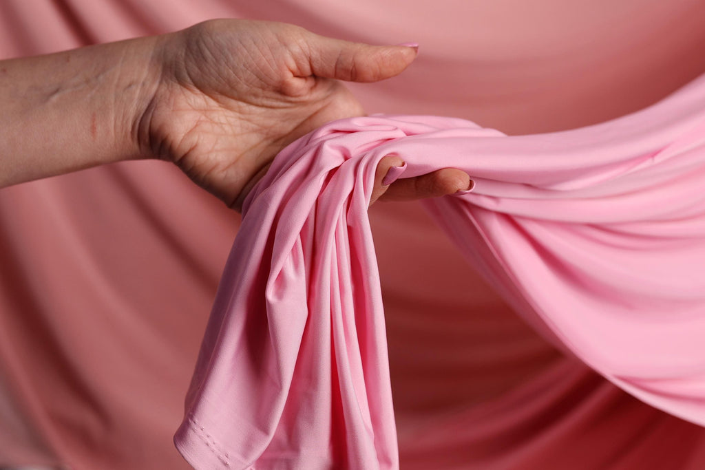 A hand is holding a pink fabric, possibly a piece of clothing, against a pink background.