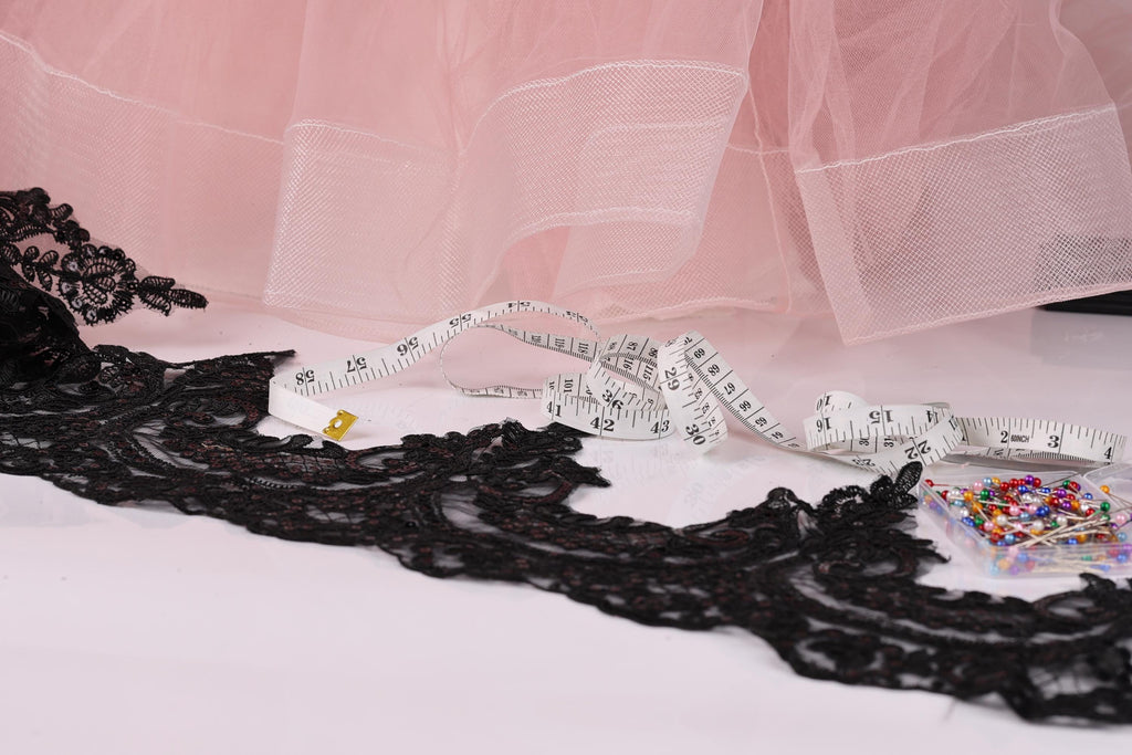 a pink tulle skirt, a black lace piece, a white measuring tape, and a small box of colorful pins.